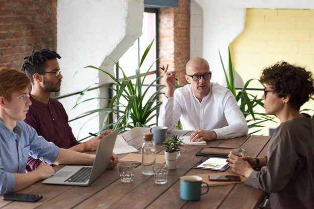 4 people sitting at a wood table talking with laptops and notepads