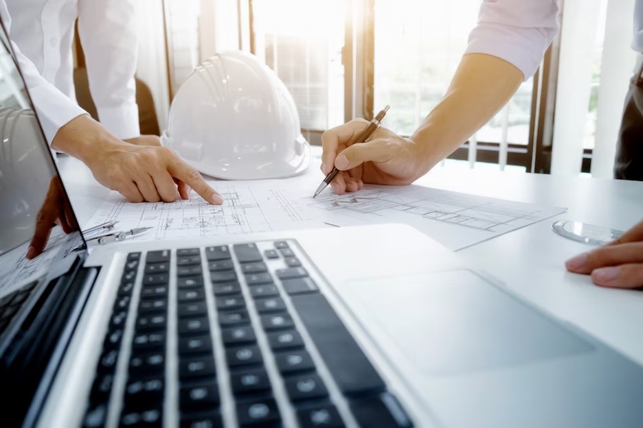 A desk with a laptop on it, construction hat, and 2 people reviewing architecture plans