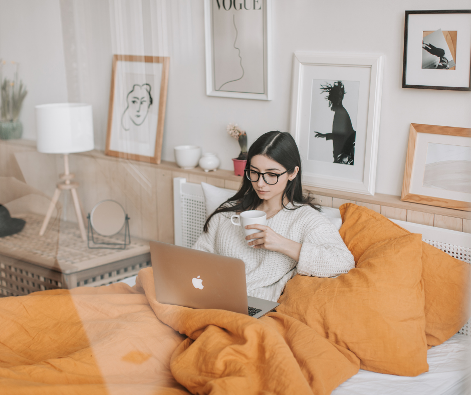 woman on a comfy bed looking at her laptop and drinking coffee